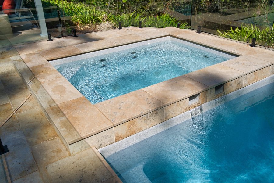 Raised spa spilling into a fibreglass pool with travertine paving
